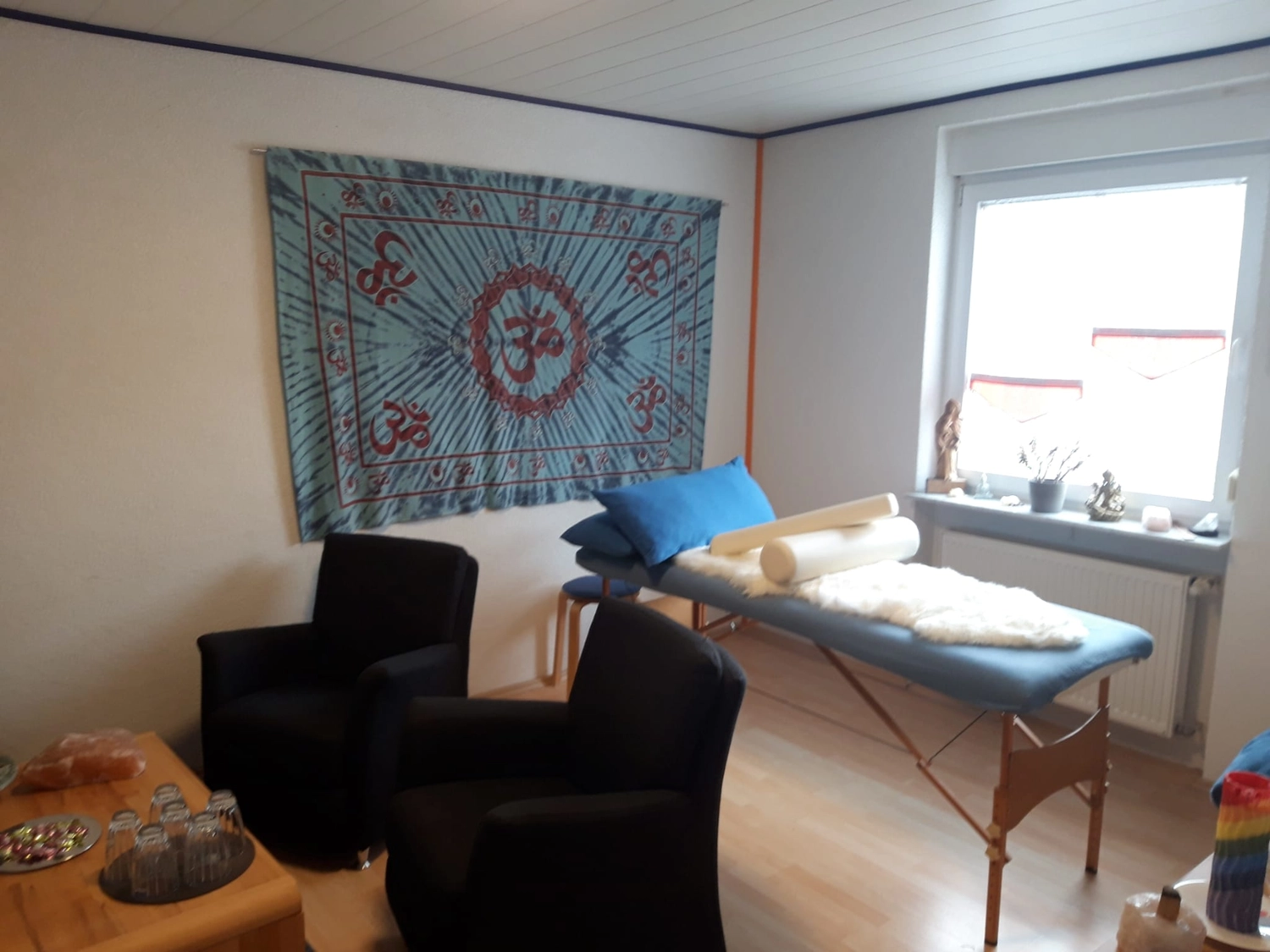 Anette Wolf – Heilpraxis – Ein ruhiger Behandlungsraum mit einer Massageliege, zwei schwarzen Stühlen und einem blauen „Om“-Wandteppich an der Wand.