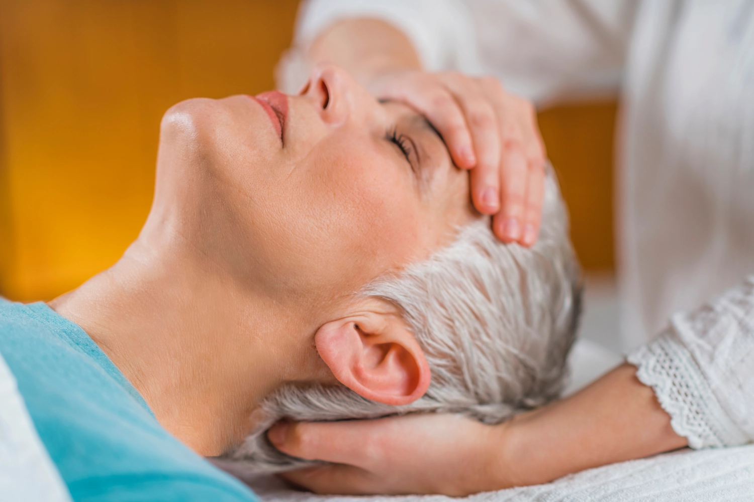 Anette Wolf – Heilpraxis – Eine Frau mit grauen Haaren liegt entspannt mit geschlossenen Augen da, während eine andere Person sanft ihren Kopf hält, wie bei einer Reiki- oder Craniosacral-Therapie-Sitzung.