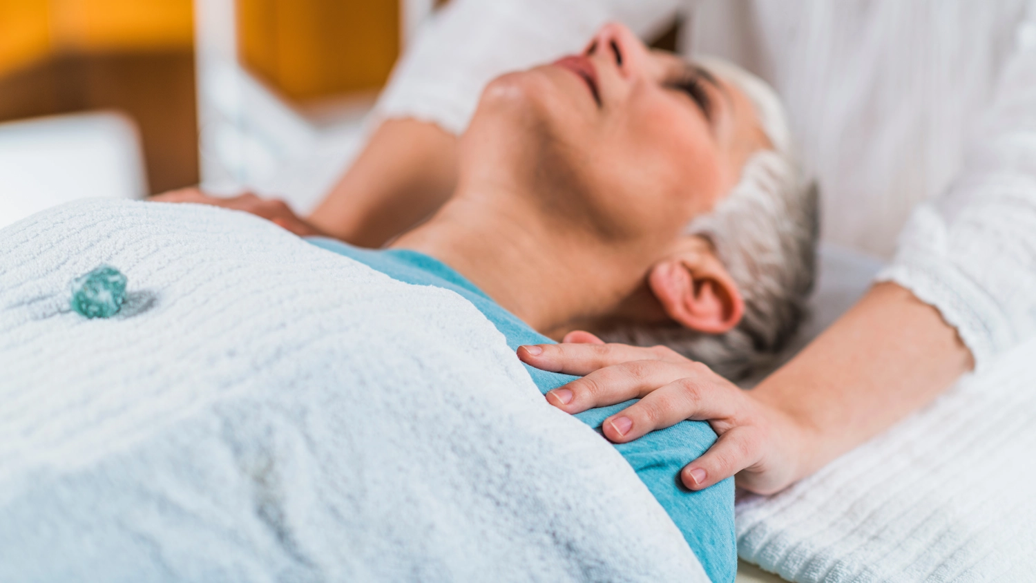 Anette Wolf – Heilpraxis – Eine Frau mit grauen Haaren liegt entspannt mit geschlossenen Augen da, während eine andere Person sanft ihren Kopf hält, wie bei einer Reiki- oder Craniosacral-Therapie-Sitzung.