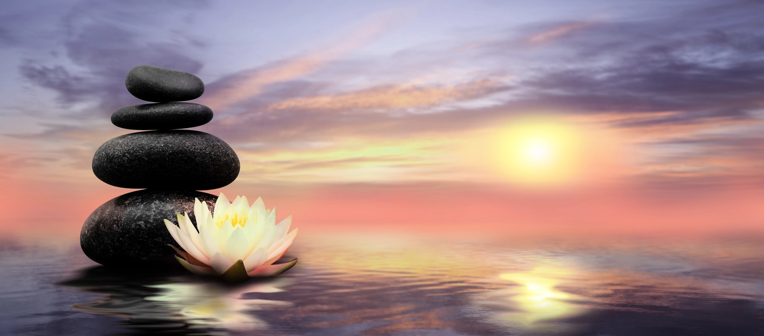 Anette Wolf – Heilpraxis – Ein Stapel aus vier glatten schwarzen Steinen und eine weiße Lotusblüte, die bei Sonnenuntergang auf ruhigem Wasser schwimmt, während am Himmel warme rosa und violette Wolken zu sehen sind.